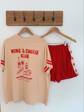 Load image into Gallery viewer, Beige t-shirt with 'Wine & Cheese Club' text and red shorts on a hanger against a white wall.