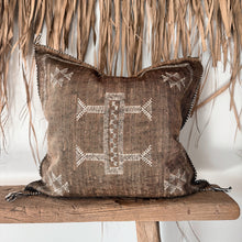 Load image into Gallery viewer, A square brown woven throw pillow with white geometric embroidery and fringed edges, resting on a rustic wooden bench in front of a dried palm leaf wall.