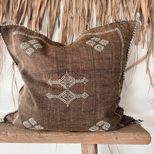 Load image into Gallery viewer, A square brown woven throw pillow with white geometric embroidered patterns, resting on a rustic wooden stool in front of a dried palm leaf wall.