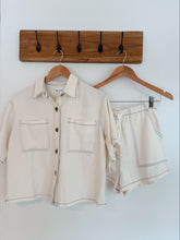 Load image into Gallery viewer, Beige shirt and shorts set on hangers against a white wall with a wooden hook board.