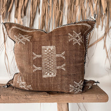 Load image into Gallery viewer, A square brown woven throw pillow with cream geometric patterns and fringed edges, displayed on a rustic wooden bench in front of a dried palm leaf wall.