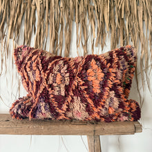 Load image into Gallery viewer, A square shaggy throw pillow with a textured, chunky knit pattern in rust red, orange, and purple, resting on a rustic wooden bench in front of a dried palm leaf wall hanging.
