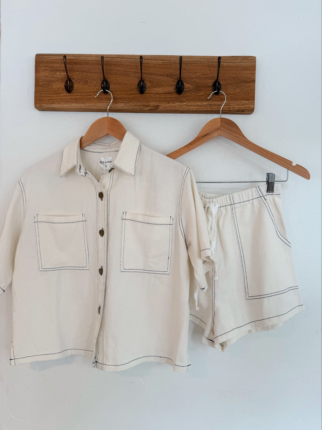 Beige shirt and shorts set on hangers against a white wall with a wooden hook board.