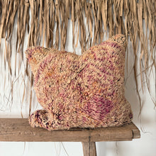 Load image into Gallery viewer, A chunky, shaggy throw pillow in a variegated blend of dusty pink, beige, and rust orange, styled on a rustic wooden bench against a backdrop of dried palm leaves.
